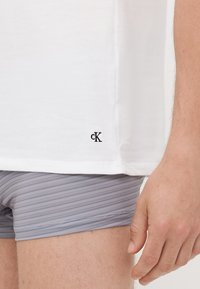 T-shirt en coton blanc avec un petit logo "cK" noir en bas, porté sur un short de boxeur gris à rayures. Manches courtes et coupe décontractée.