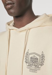 Sudadera con capucha color crema con cordón, que presenta un logo gráfico negro de un edificio y detalles de laurel en el pecho. Tela suave y casual.