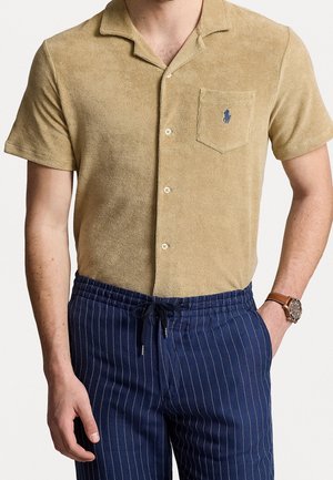 Homme portant une chemise beige à manches courtes avec poche poitrine et un pantalon bleu marine à fines rayures avec ceinture à cordon.