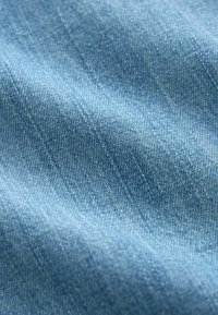 Close-up of textured light blue denim fabric with visible diagonal weave and soft folds creating shadows and highlights.