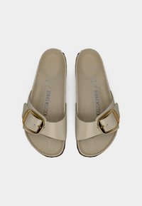 Ζευγάρι μπεζ σαγιονάρες Birkenstock με μεγάλες χρυσές αγκράφες και εσοχές για τα πέλματα, εμφανιζόμενες από πάνω σε λευκό φόντο.