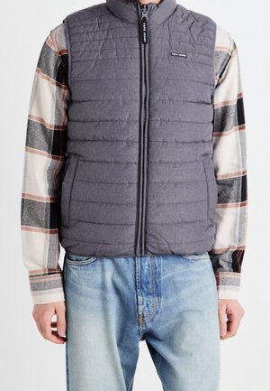 Muž v šedé prošívané vestě přes béžovou a černou károvanou košili s dlouhým rukávem a světlými modrými džínami, stojící před neutrálním pozadím.