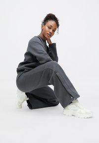 Conjunto de fato de treino cinza com um ajuste relaxado, apresentando gola redonda e detalhes laterais. Combinado com ténis brancos robustos, destacando um design moderno.