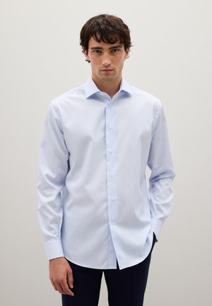 Joven vestido con una camisa de vestir de manga larga azul claro y pantalones oscuros, de pie contra un fondo neutro y liso.