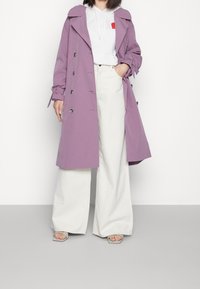 Lavendel trenchcoat med dubbelknäppt design, stor krage och knyt detaljer. Kombinerad med en vit hoodie och vida ljusa byxor.