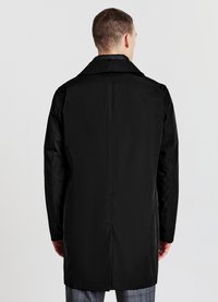 Homme portant un long manteau noir avec un col, se tenant de dos contre un fond blanc uni.