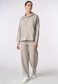 Felpa leggera color beige chiaro con zip e pantaloni ampi coordinati. Tessuto liscio con una texture morbida. Logo evidente sul petto. Sneakers bianche.