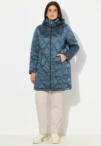 Vadderad, huva jacka i mörkblått med en glansig finish. Har en framsida med dragkedja, sidofickor och knälång design. Dekorerad med diamantmönster.