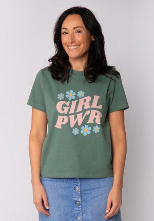 Femme aux cheveux bruns ondulés souriant, portant un t-shirt vert "GIRL PWR" avec des fleurs bleues et une jupe en jean bleue boutonnée.