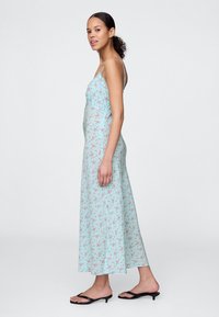 Modra obleka maxi s cvetličnim potiskom, tankimi naramnicami, oprijetim zgornjim delom in rahlo razširjenim robom, kombinirana s črnimi ravnimi sandali.