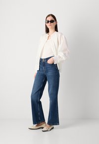 White, oversized button-up shirt, fitted white tank top, high-waisted blue flared jeans, black sunglasses, cream flat shoes with stud details.