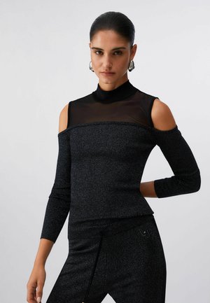 Donna con capelli tirati all’indietro indossa un top nero con spalle scoperte e scollo trasparente abbinato a pantaloni strutturati su sfondo chiaro uniforme.