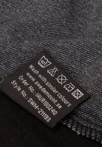 Textiellabel met zorgsymbolen op een zwarte stof achtergrond. Textuur van grijze stof zichtbaar in het bovenste gedeelte.