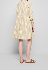 Robe rayée jaune et grise, longueur genou avec une conception en étages, manches trois-quarts avec liens, associée à des sandales à glissière marron.