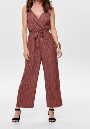 Woman wearing sleeveless maroon patterned wide-leg jumpsuit with V-neck and waist tie, paired with black ankle-strap heels and gold bracelet.