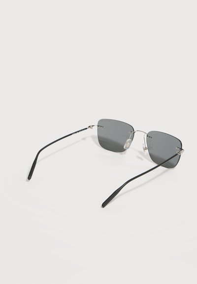 Κομψά γυαλιά ηλίου χωρίς σκελετό με σκούρους ορθογώνιους φακούς και λεπτά μαύρα μπράτσα που ακουμπούν σε λευκή επιφάνεια.