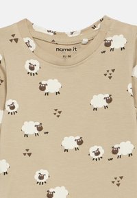 Bežinė kūdikio bodži su pakartotiniais baltais avinėlių raštais ir mažomis rudos trikampių grupėmis, dydis EU 56, apvalus iškirpimas su siuvinėjimo detalėmis.