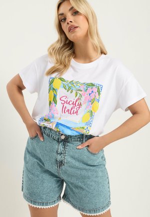 Ung kvinde med blondt hår iført hvid Sicily Italia T-shirt og højtaljede acid-wash denimshorts med blondekant.