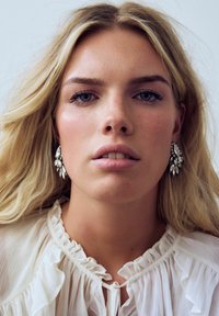 Silver-tone floral earrings with clear rhinestones, worn by a model with long, wavy blonde hair. The blouse is light, with ruffled details.