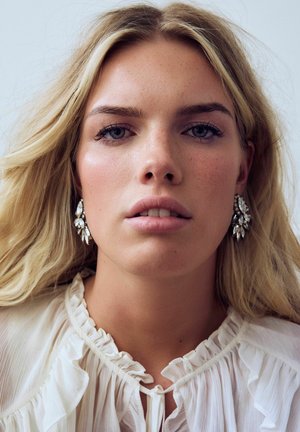 Silberfarbene Blumenohringe mit klaren Strasssteinen, getragen von einem Model mit langen, welligen blonden Haaren. Die Bluse ist leicht und hat Rüschendetails.