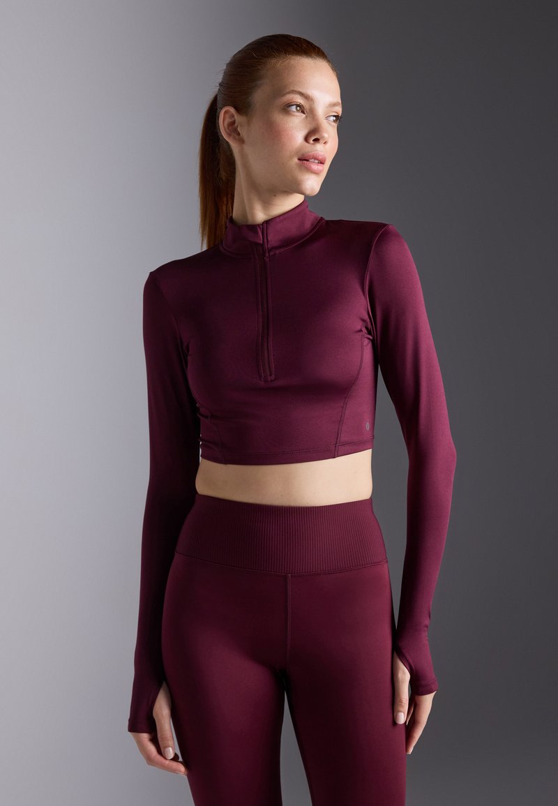 Bordo atletski crop top s dugim rukavima, visokim ovratnikom i prednjim patentnim zatvaračem; u kombinaciji s odgovarajućim tajicama visokog struka, glatka tekstura tkanine.