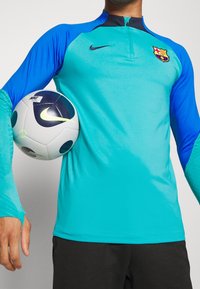 Nike långärmad sporttröja i teal och blått, med en kvartdragkedja, Barca-logotyp och texturerade detaljer. Håller en svart och vit fotboll.