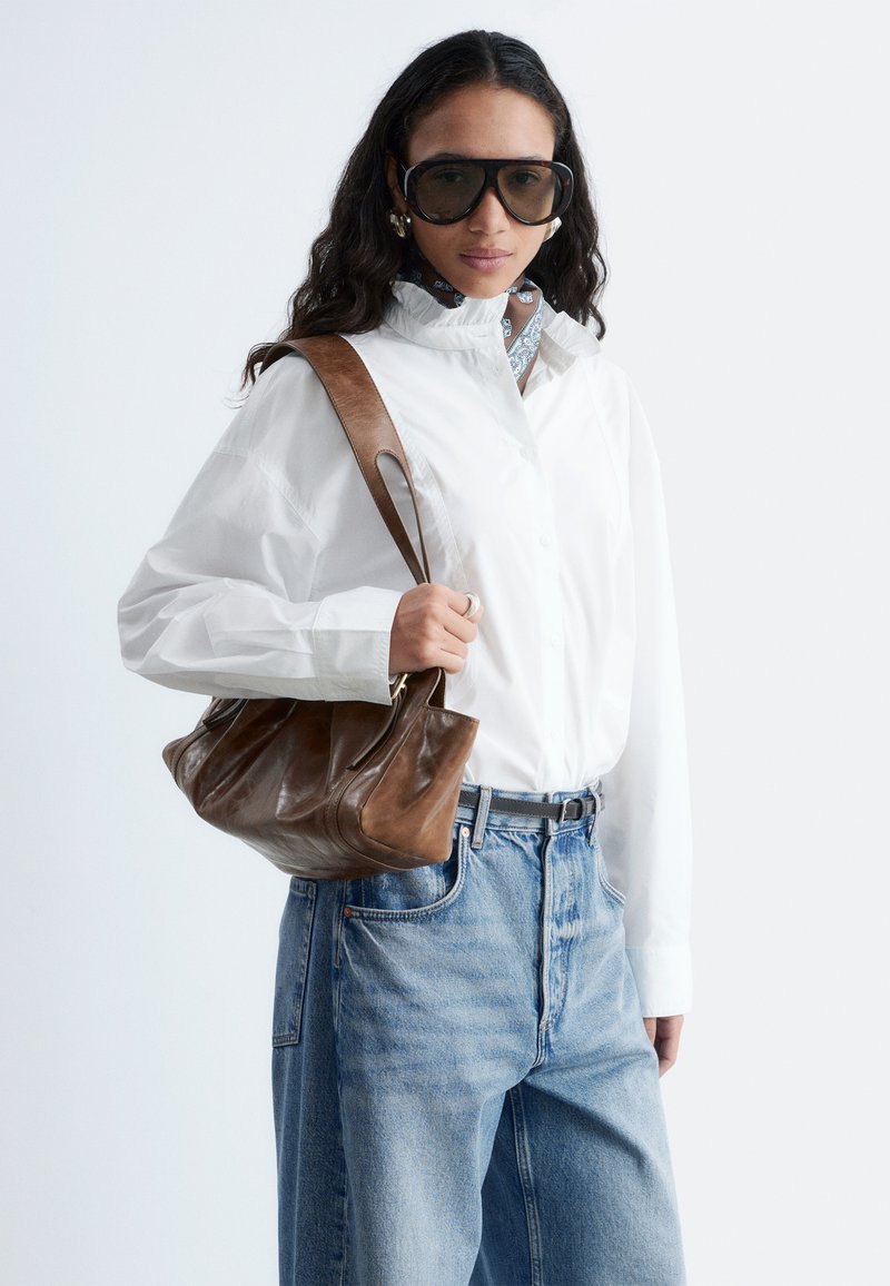 Femme aux longs cheveux bruns portant de grandes lunettes de soleil, une chemise blanche boutonnée, un jean bleu, un sac à épaule en cuir marron et un foulard à motifs autour du cou.