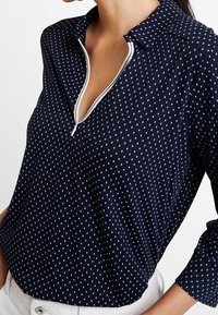 Marineblaue Bluse mit einem kragenlosen Ausschnitt, halbem Reißverschluss und weißen Punkten. Weicher Stoff, lockere Passform mit umgeschlagenen Ärmeln.
