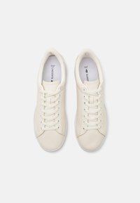 Lacoste-tennarit off-white nahasta, pyöreällä kärjellä, nauhakiinnityksellä ja teksturoiduilla yksityiskohdilla. Varustettu kontrastivärisellä valkoisella pohjalla.