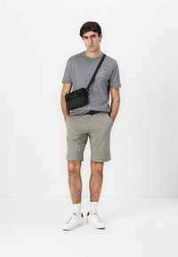 T-shirt grise à manches courtes, shorts vert clair, sac bandoulière noir, baskets blanches et chaussettes blanches. Design minimaliste et couleurs neutres.