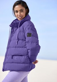 Purple puffer jacket with a high collar, zippered front, and elastic drawstrings. Features a black logo patch on the left sleeve and front pocket.