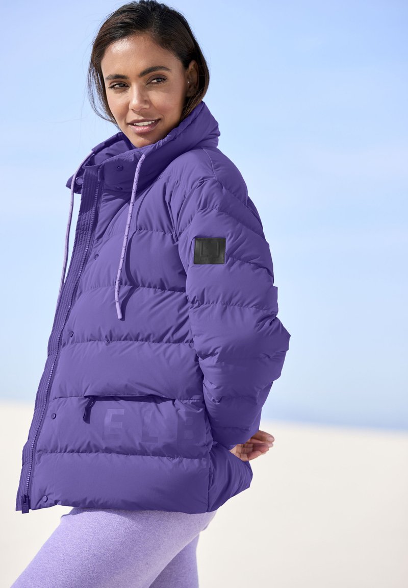 Purple puffer jacket with a high collar, zippered front, and elastic drawstrings. Features a black logo patch on the left sleeve and front pocket.