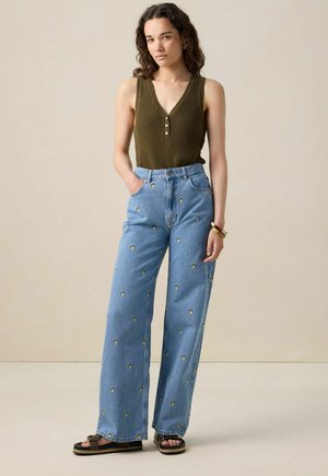 Woman wearing sleeveless olive green ribbed top with buttons, light blue wide-leg jeans with small yellow flower prints, and black sandals.