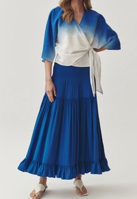 Blauwe ombre wikkel top met brede mouwen en een witte strik, gecombineerd met een lange, gelaagde blauwe rok met een gerimpelde zoom. Platte sandalen.