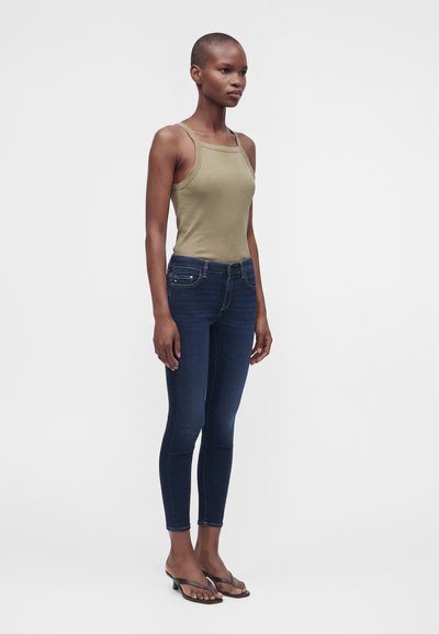 Γυναίκα στέκεται φορώντας ελιάς πράσινη μπλούζα χωρίς μανίκια, σκούρο μπλε στενά τζιν και μαύρα πέδιλα με τακούνι και ανοιχτά δάχτυλα, σε λευκό φόντο.