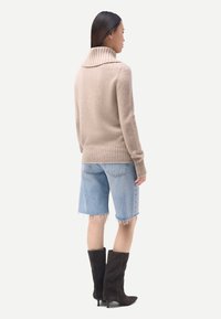 Femme portant un pull à col roulé beige, un short en denim bleu clair avec des ourlets effilochés, et des bottes noires montantes à talons, debout de profil.