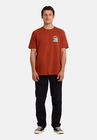 Rdzawoczerwony bawełniany t-shirt z okrągłym dekoltem, krótkimi rękawami oraz grafiką słońca i fal na lewej piersi. Połączony z czarnymi spodniami.