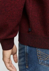 Pulover texturat roșu și negru cu o margine ribată. O etichetă mică neagră este vizibilă la marginea de jos. Împreună cu blugi din denim albastru.