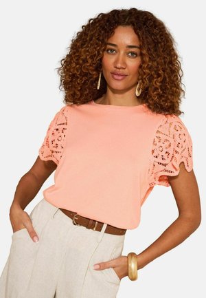 Femeie cu păr creț maro poartă un top piersică cu mâneci din dantelă, pantaloni bej cu talie înaltă, curea maro, brățară aurie și cercei cerc.