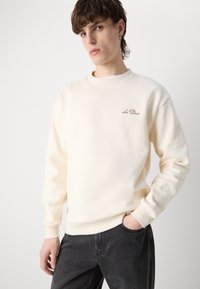 Sudadera color crema con cuello redondo, puños acanalados y un sutil logo bordado en el pecho. Tejido suave y texturizado, corte relajado.