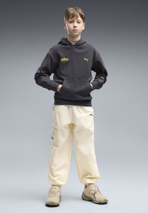 Garçon debout les mains dans la poche avant, portant un sweat à capuche noir Pokémon-Puma, un pantalon cargo crème et des baskets beige sur fond gris.