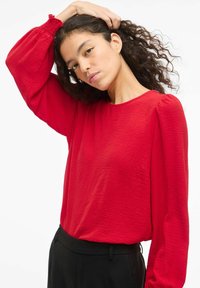 Blusa roja de manga larga con tejido texturizado, escote redondo y mangas abullonadas, combinada con pantalones negros frente a un fondo neutro.