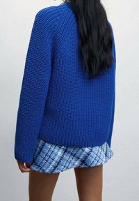 Femme aux longs cheveux noirs portant un gros pull en tricot bleu et une jupe plissée bleue, debout devant un fond clair uni.