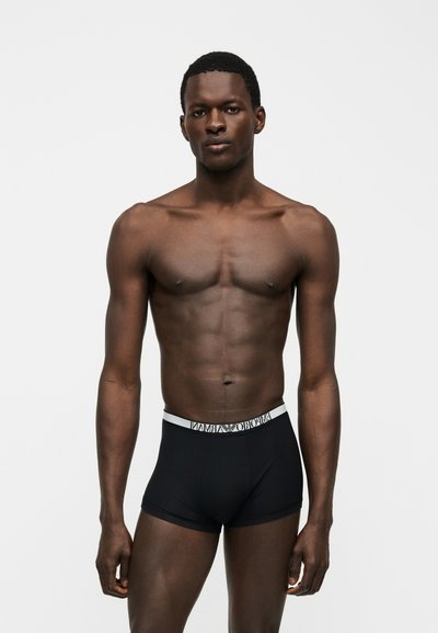 Άνδρας μοντέλο με καλογυμνασμένο σώμα που φοράει μαύρα boxer Emporio Armani σε λευκό φόντο.