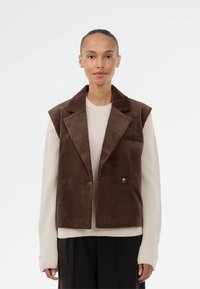 Brązowy żakiet bez rękawów z korduroyu, z wyciętymi klapami, podwójnym zapięciem na guziki i boczną kieszenią, nałożony na jasny sweter z dziewiarki.