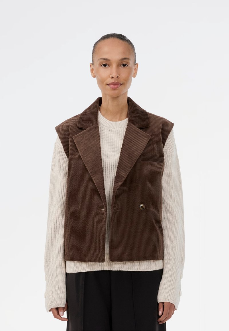Brązowy żakiet bez rękawów z korduroyu, z wyciętymi klapami, podwójnym zapięciem na guziki i boczną kieszenią, nałożony na jasny sweter z dziewiarki.