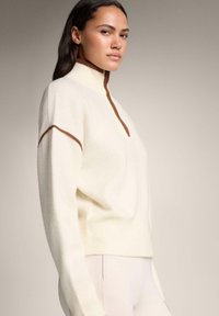 Džemper boje kreme s visokim ovratnikom, smeđim rubom i rebrastim rubom. Meka tekstura, opušten kroj i bočni prorezi za dodatni detalj.