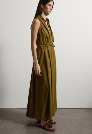 Vrouw draagt een mouwloze olijfgroene maxi-jurk met riem, bruine gevlochten sandalen en een rode kralenketting, staand op een grijze vloer.