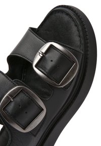 Sandalia de cuero negro con dos correas anchas, que presenta grandes hebillas metálicas plates. La suela es plana y texturizada para proporcionar agarre.