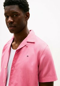 Camicia rosa a maniche corte con una consistenza morbida, design con bottoni e un piccolo logo sul lato sinistro del petto. Canottiera bianca visibile sotto.
